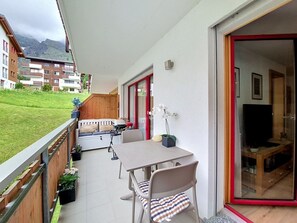Ein Balkon mit Tisch und Stühlen, einer Couch, einem Fernseher und Blick auf Gebäude und Berge.