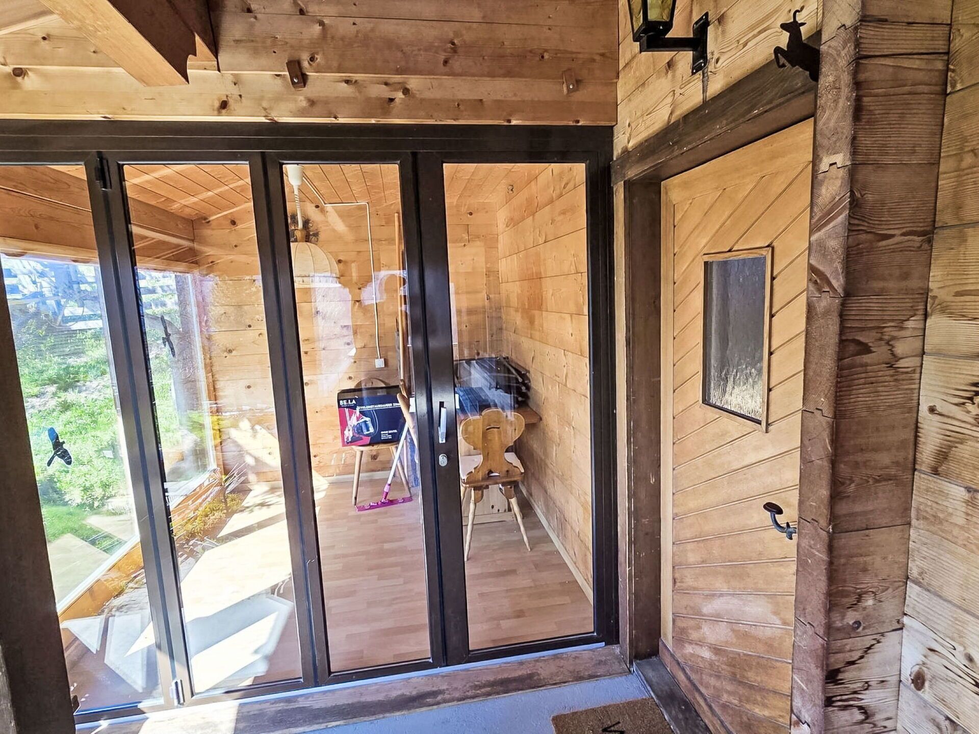 Un interior de madera con un puertas correderas de vidrio que dan a un balcón, una pequeña mesa con una silla y una lámpara fijada a la pared.