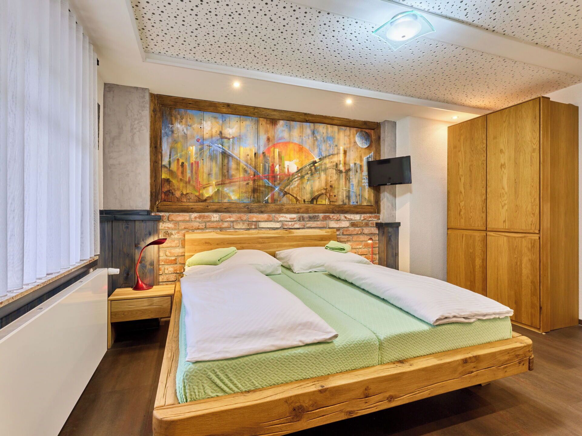 A bedroom with a wooden bed, a brick wall, a painting, a TV, and a wardrobe.