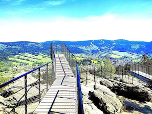 Eine Holz-Hängebrücke mit Metallgeländern, die zu einer gebirgigen Landschaft mit tiefen Tälern und blauem Himmel führt.
