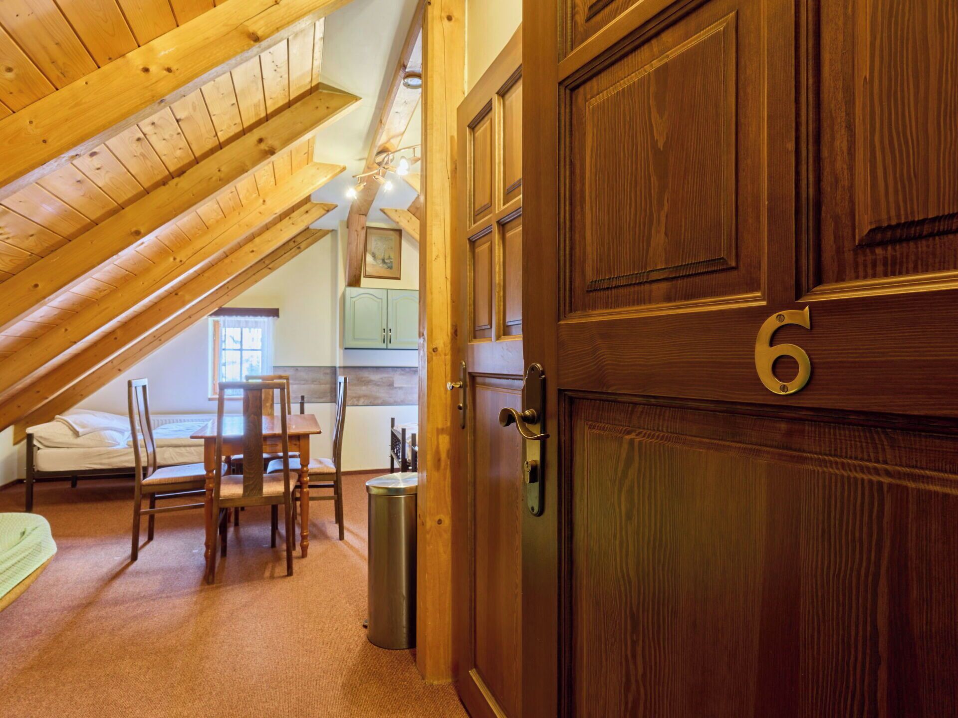 A room with a sloped ceiling, wooden door with a number, a bed, a table with chairs, and a trash bin.