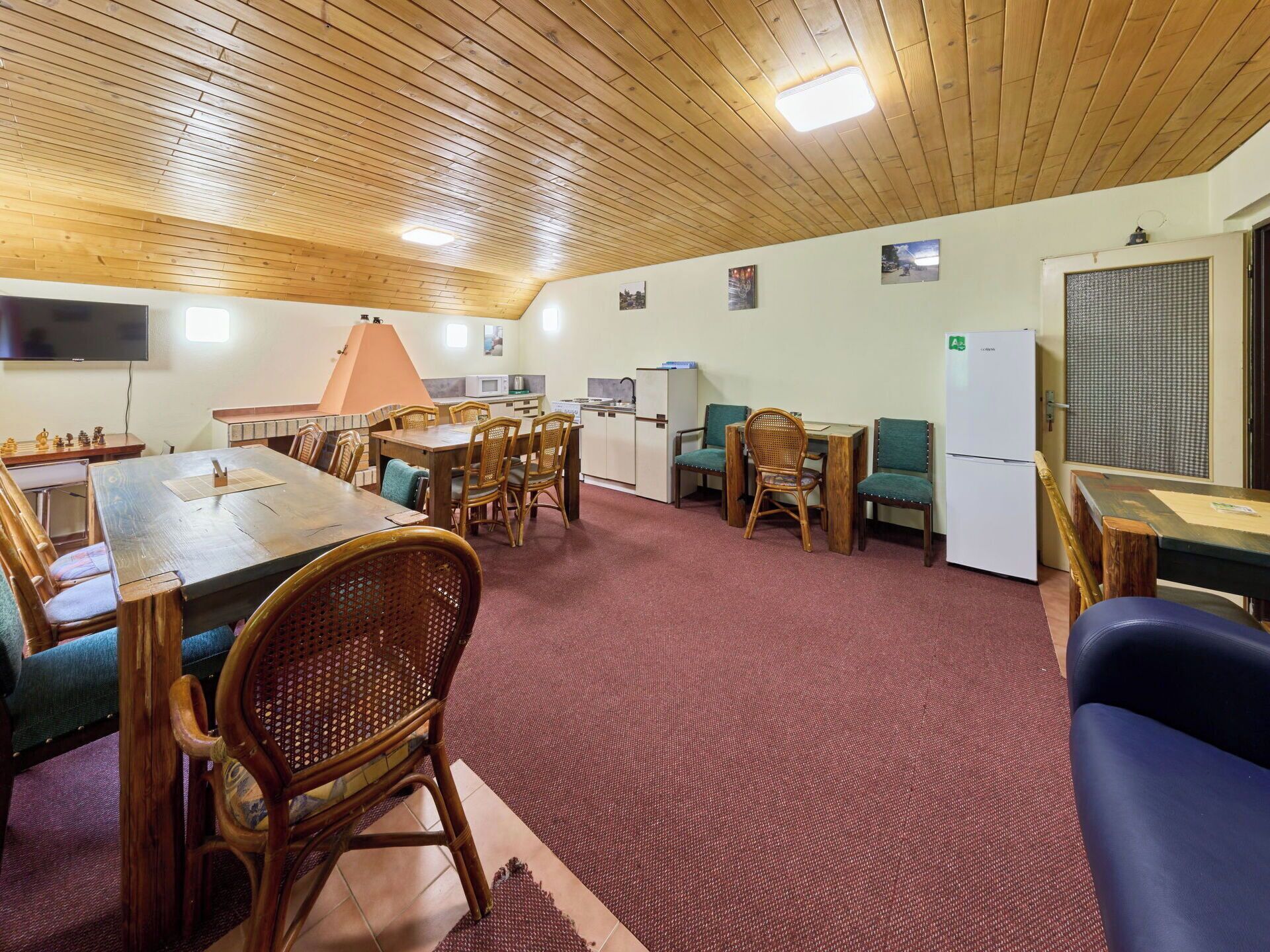 Une pièce avec un plafond en bois, une table à manger avec des chaises, un téléviseur, un réfrigérateur et une fenêtre avec des stores.