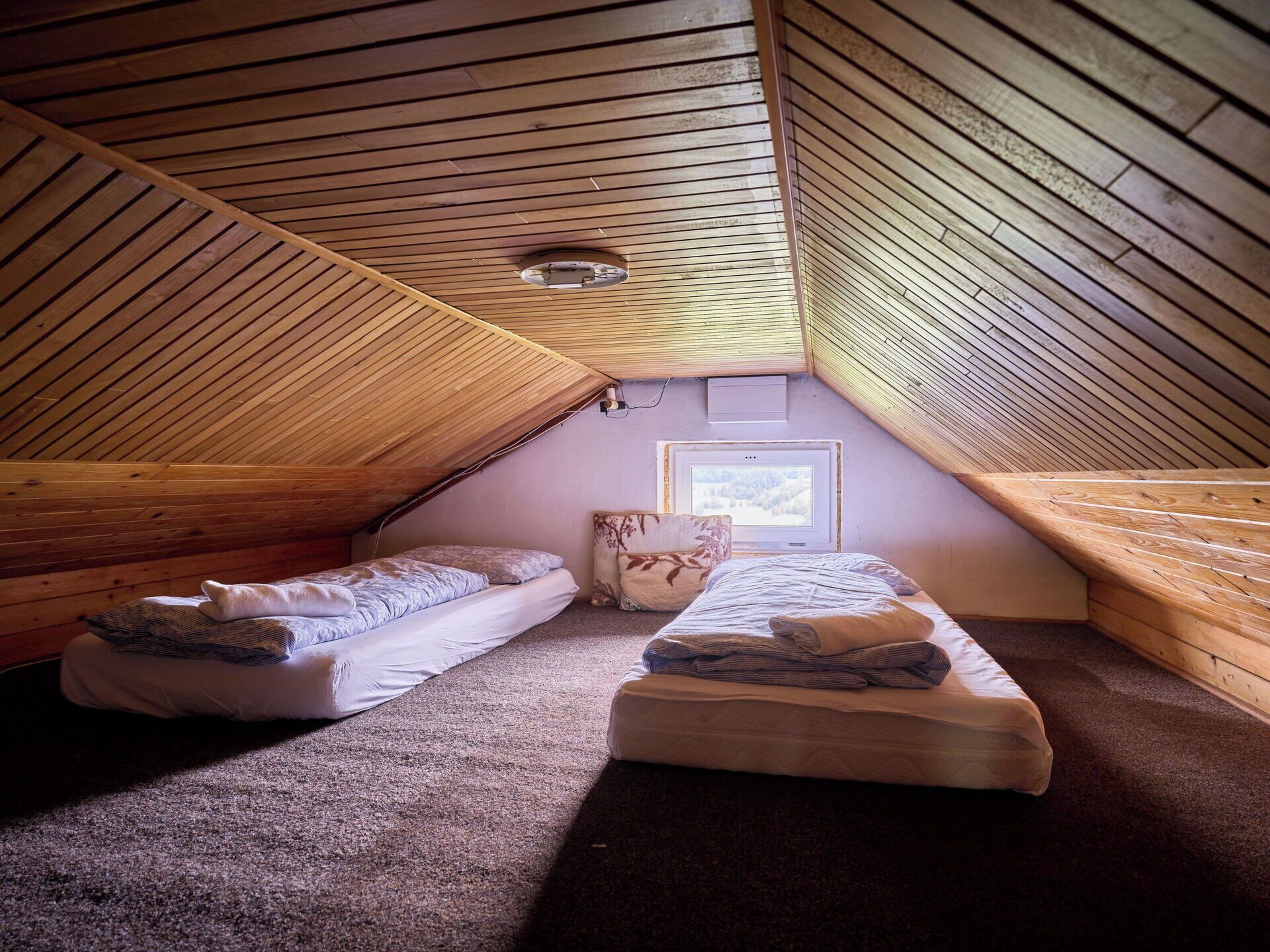 Ein kleines Schlafzimmer im Dachgeschoss mit geneigter Holzbalkendecke, zwei Betten, einem Fenster mit Blick und einem Wandheizkörper.