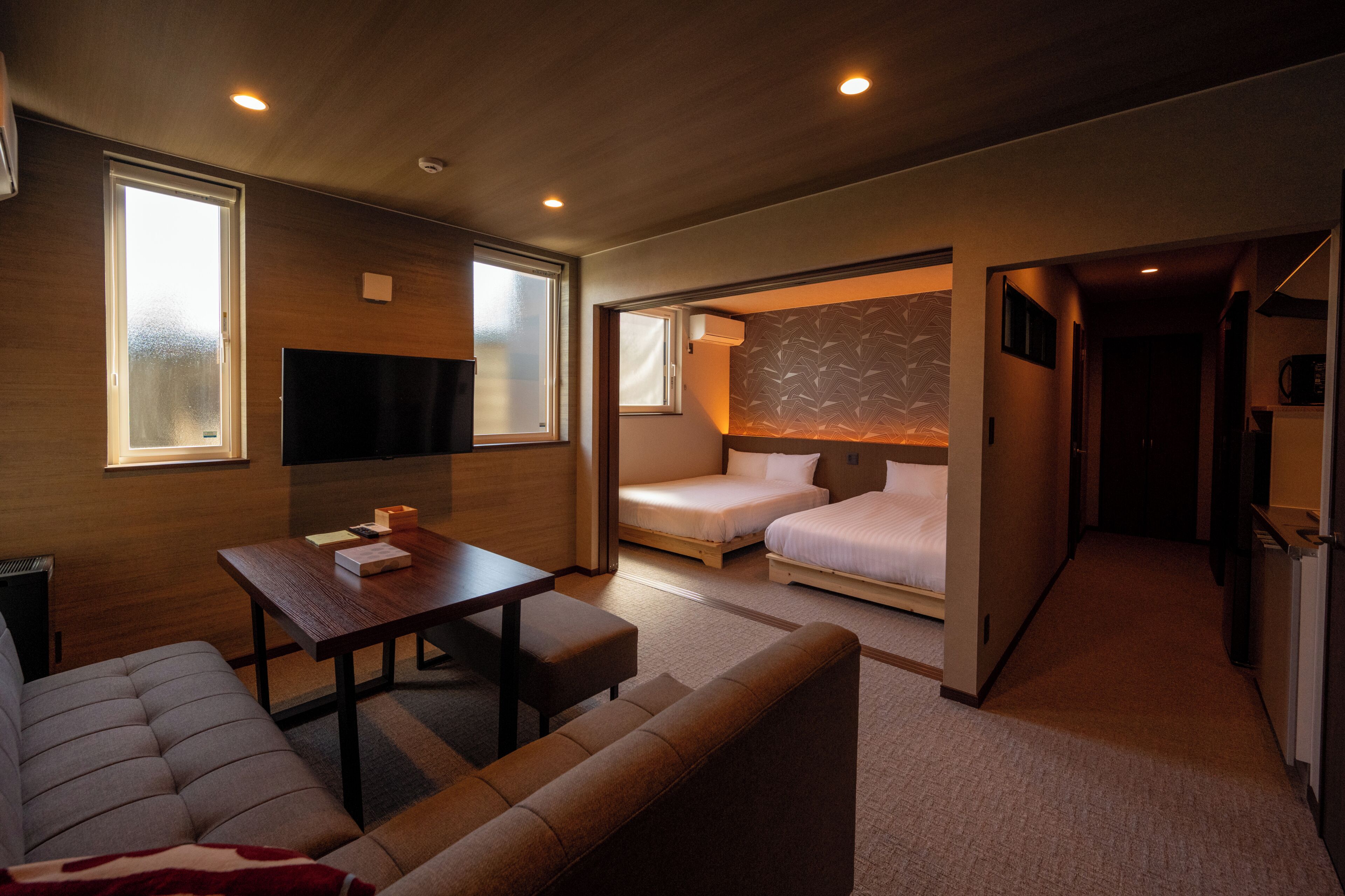 A hotel room with a sofa, a table, two beds, a TV, and a kitchen area.