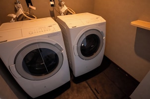 Dos lavadoras de carga frontal en un lavadero con un estante de madera y un enchufe eléctrico en la pared.