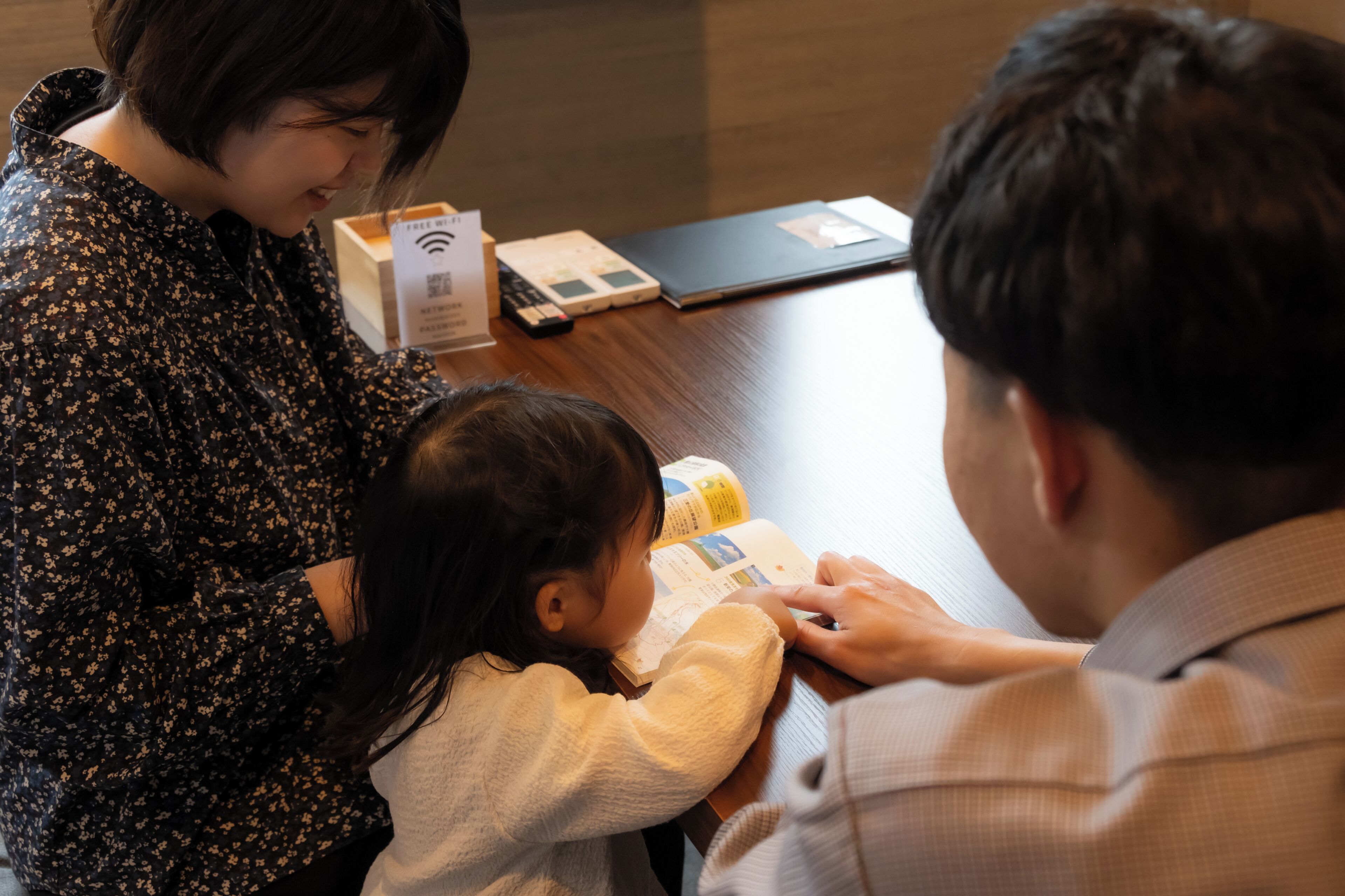 A family of three at a wooden table with a laptop, a book, and a box.
