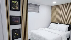 Ein modernes Schlafzimmer mit einem Bett, einem an der Wand befestigten Bilderrahmen, einer Deckenleuchte und einem Fenster mit Jalousien.