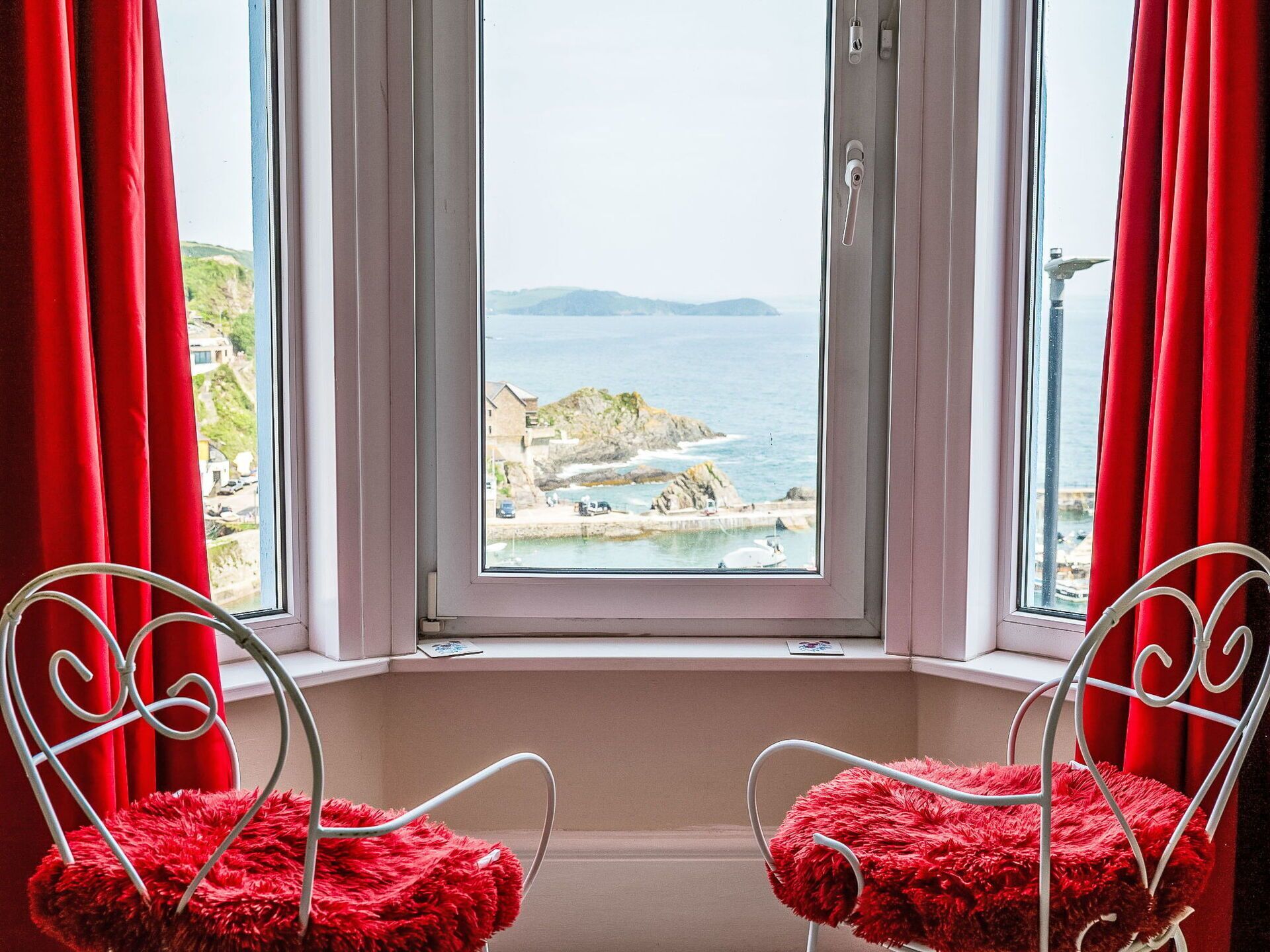 Una habitación con un ventanal grande con vistas a la costa, dos sillas metálicas con cojines rojos y una puerta blanca.