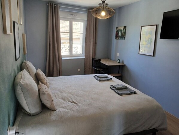 Un dormitorio con cama, almohadas, un escritorio, una silla, una ventana con cortinas, un televisor montado en la pared y obras de arte.