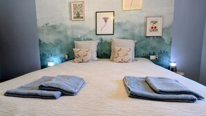 Ein ordentlich bezogenes Bett mit zwei gefalteten Handtüchern, eine Wand mit gerahmten Bildern und ein kleiner Tisch mit einer Lampe.