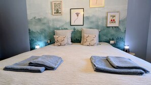 A neatly made bed with two folded towels, a wall with framed artwork, and a small table with a lamp. - Quiet 4-person house in a village; pets allowed (Mognéville)