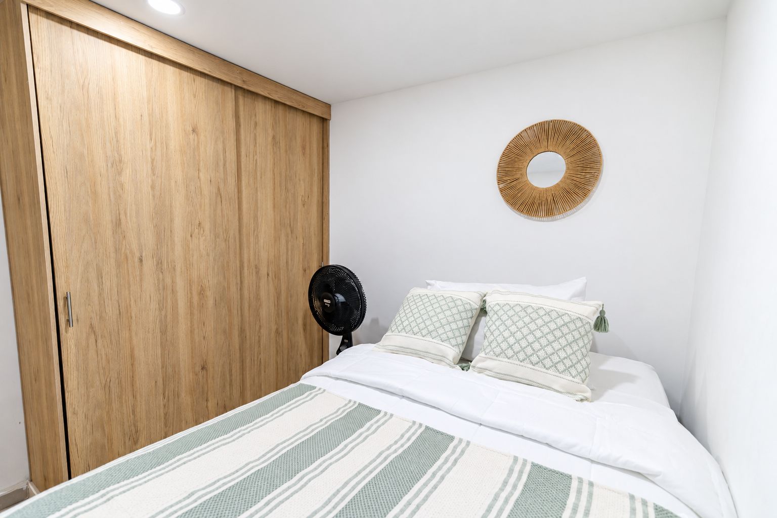 Un dormitorio con un armario de madera, una cama con ropa de cama blanca, un adorno circular en la pared y un ventilador de pie.
