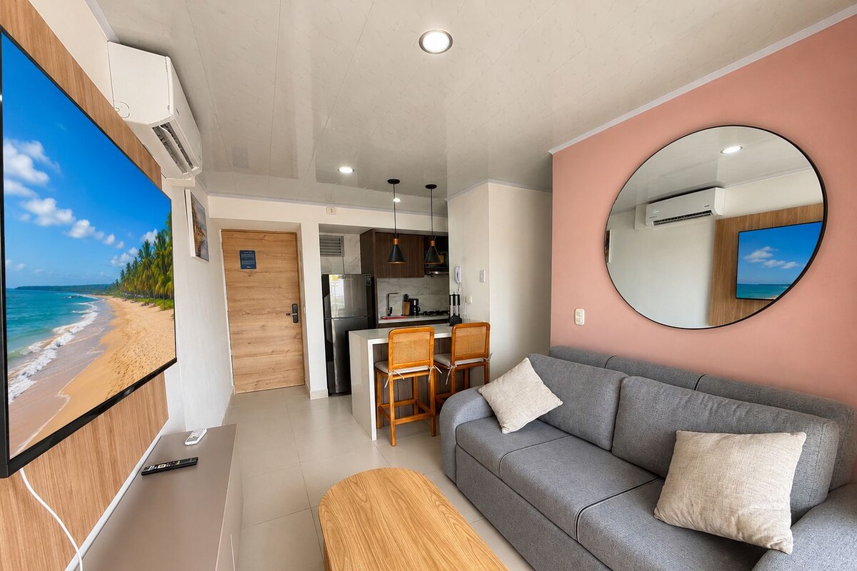Una sala de estar moderna con un sofá gris, una mesa de centro de madera y un televisor de pantalla plana grande mostrando una escena de playa.