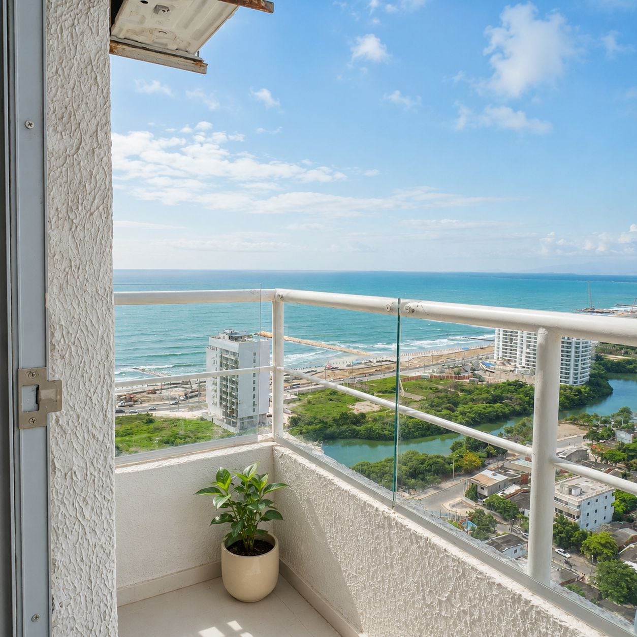 A balcony with a potted plant, overlooking a coastal cityscape with buildings and a beach.