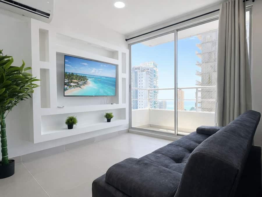 Una sala moderna con un televisor de pantalla plana grande, un sofá gris, una planta en maceta y un balcón con vista a la ciudad.