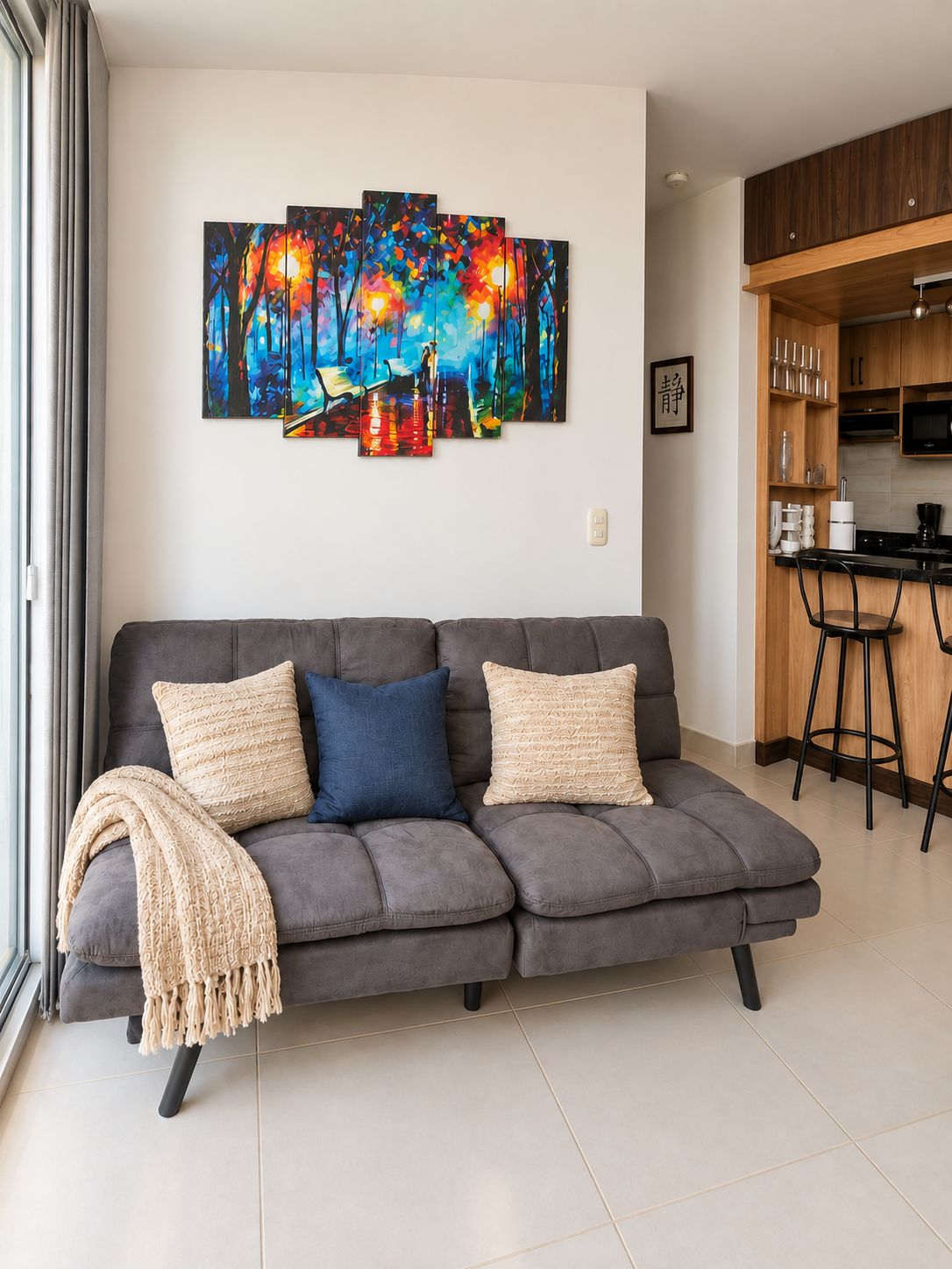 A modern living room with a grey sofa, a colorful abstract painting, and a kitchen area with wooden cabinets and a black countertop.