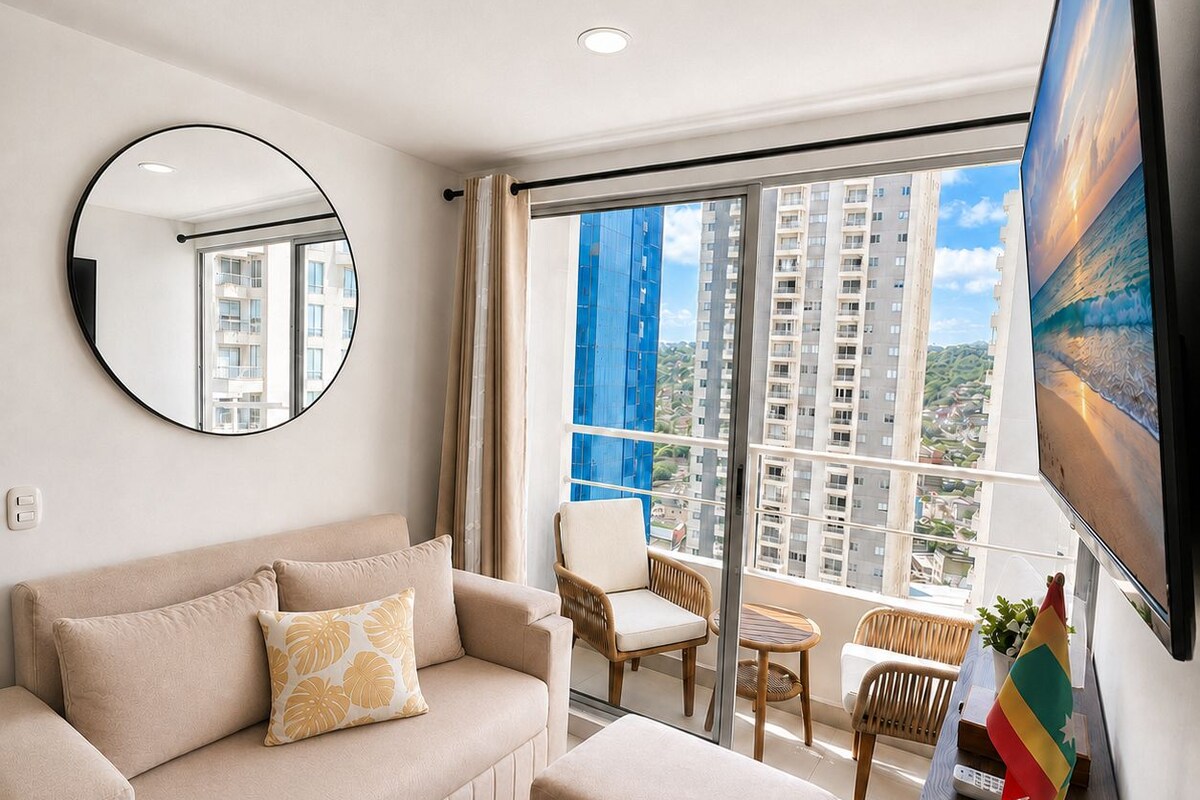 Una sala de estar moderna con un sofá, un reposapiés y una mesa de centro. Hay un televisor montado en la pared, un espejo redondo y una ventana con vistas a edificios.