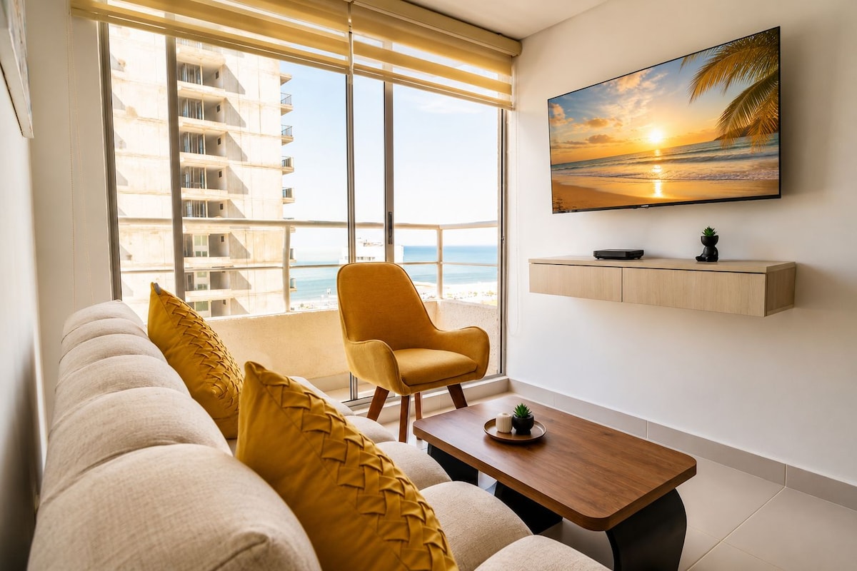 A modern living room with a sofa, armchair, and coffee table, a TV mounted on the wall, and a view of the ocean through large windows.