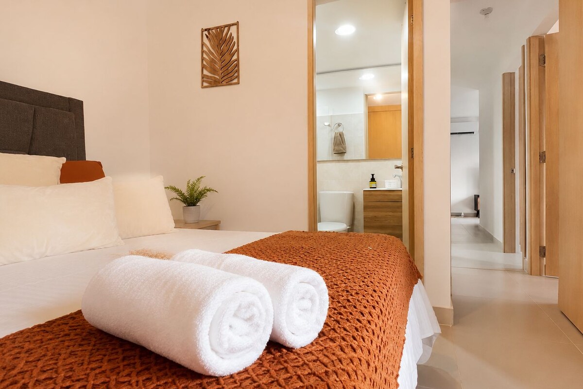Habitación de hotel con cama, mesita de noche con planta, espejo y baño visible a través de una puerta abierta.