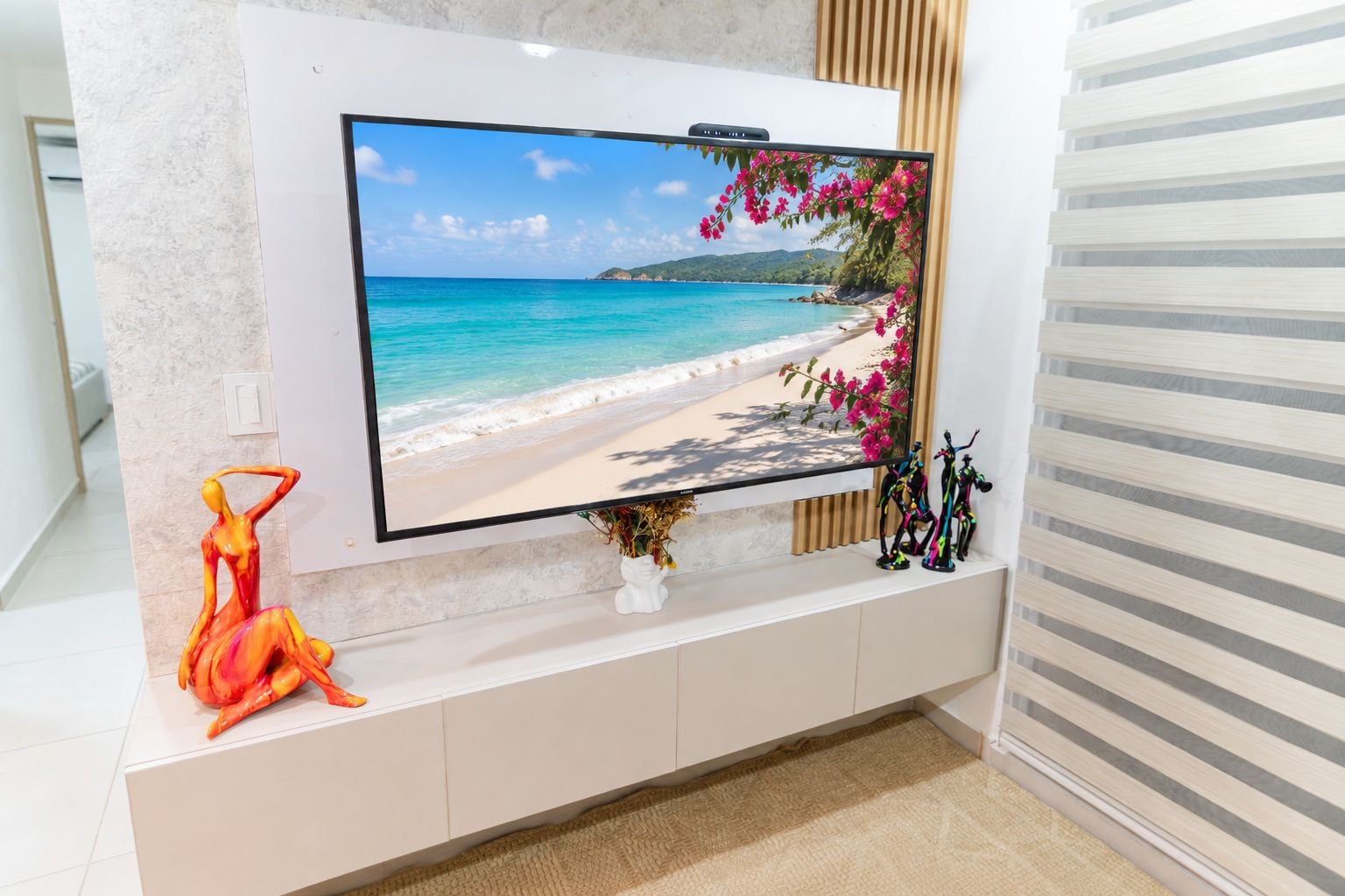 A flat-screen TV mounted on a wall with a beach scene, a white vase with flowers, and a sculpture on a shelf below.