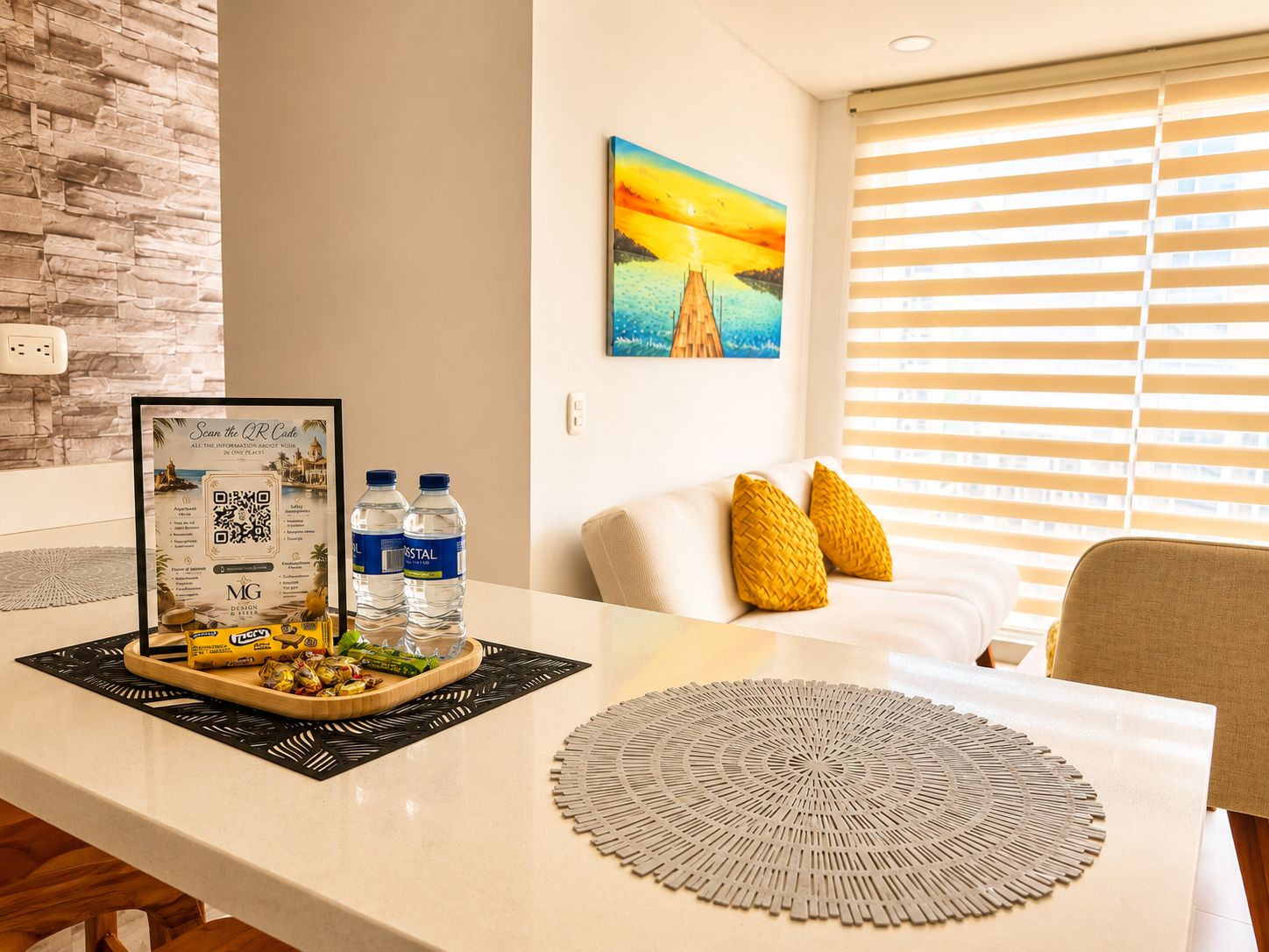 A hotel room with a bed, a bedside table with water bottles, a framed QR code, and a painting of a sunset.
