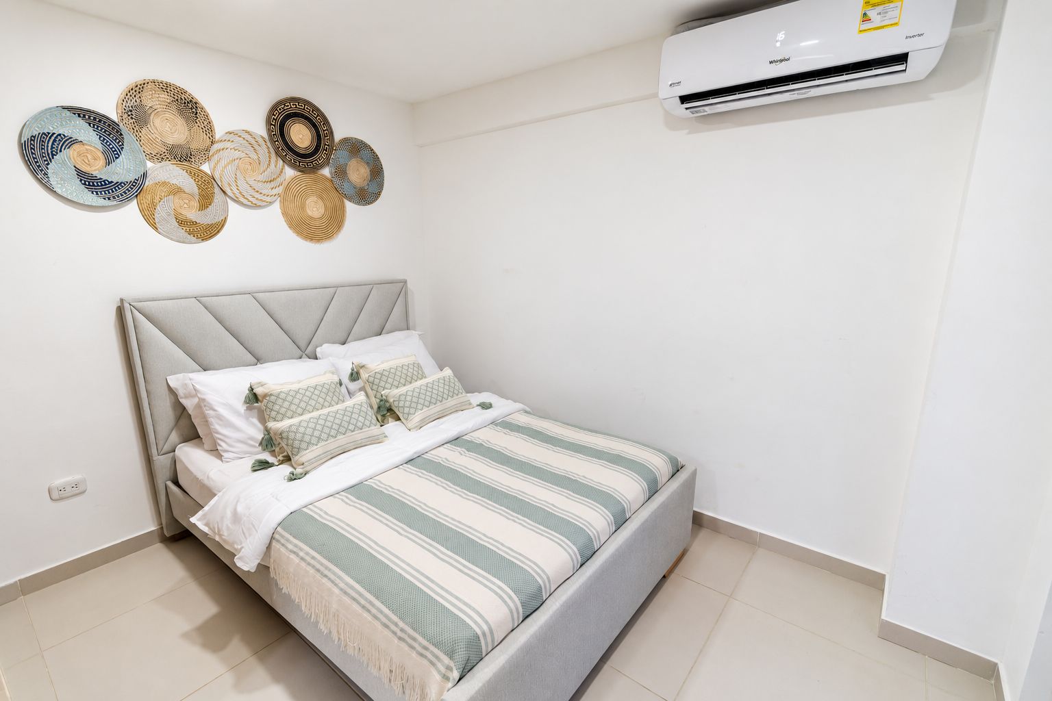 Une chambre à coucher avec un lit, des oreillers, un climatiseur fixé au mur et des œuvres d’art décoratives accrochées au mur.