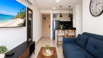 Una sala de estar moderna con un televisor de pantalla plana grande, un sofá azul, una mesa de centro de madera y una zona de cocina con refrigerador y juego de comedor.