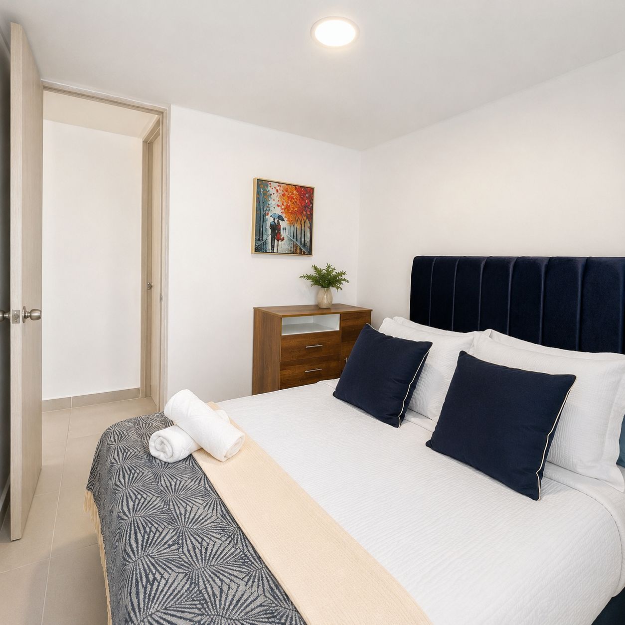A bedroom with a bed, pillows, a bedside table with a plant, and a painting on the wall.