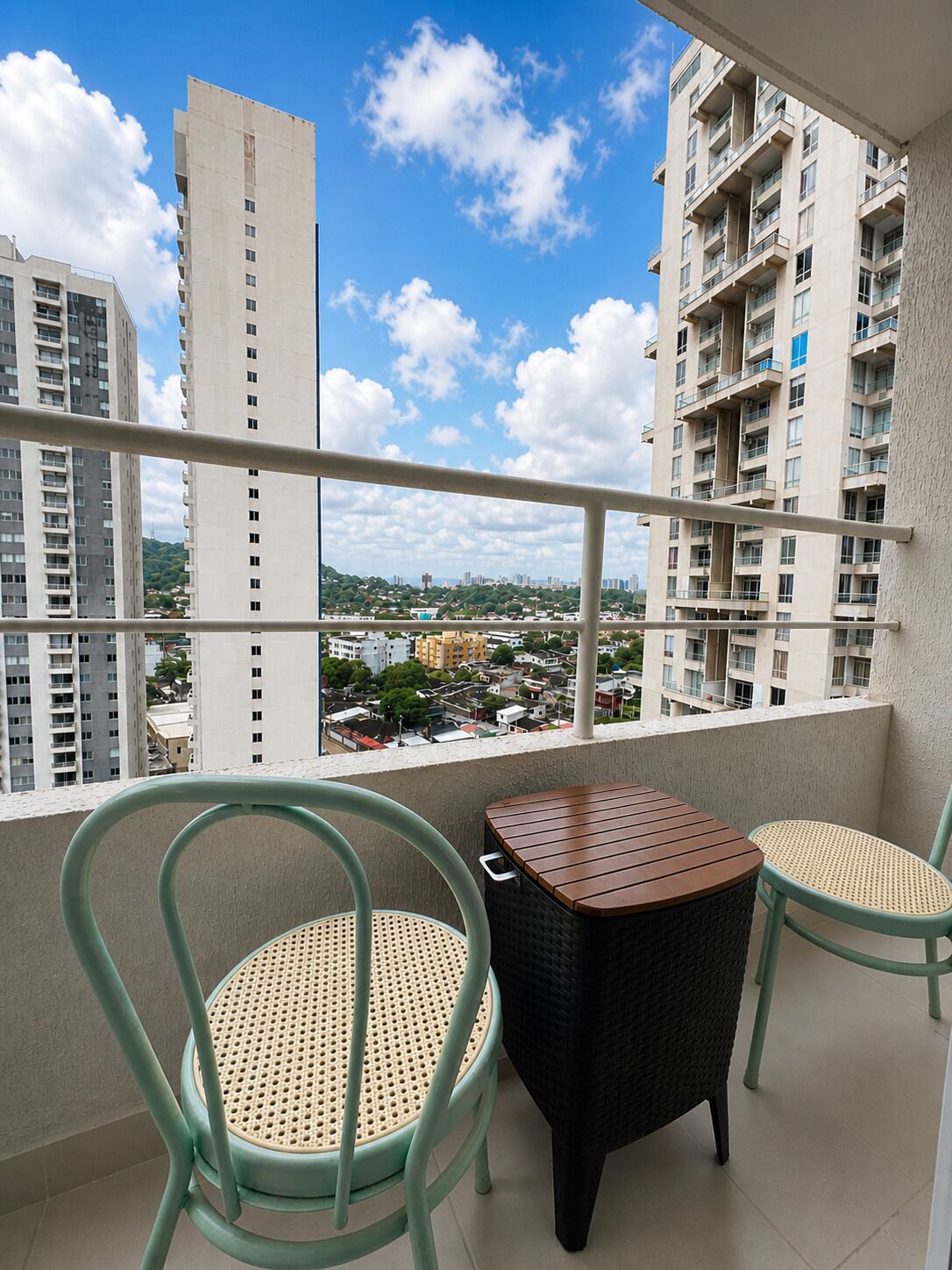 A balcony with two wicker chairs and a small table, overlooking a cityscape with high-rise buildings and a clear sky.