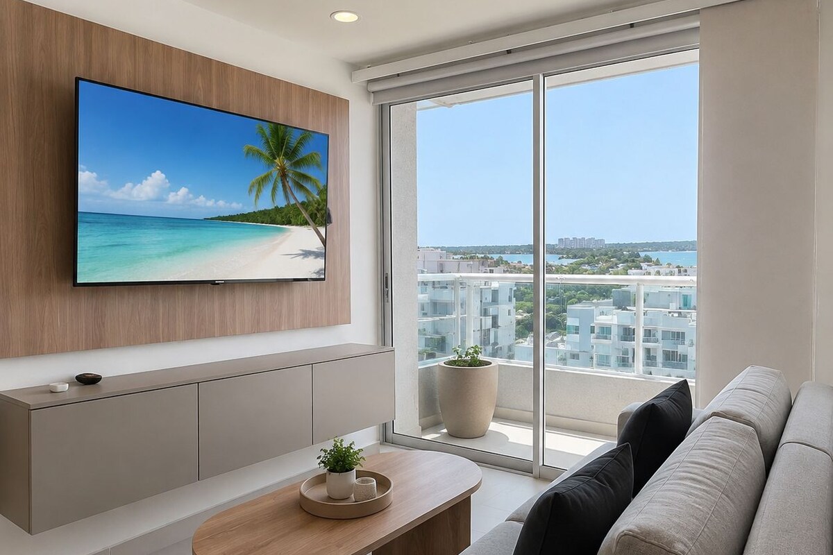 Una sala de estar moderna con un televisor grande, un sofá, una mesa de centro y vistas a la playa y al paisaje urbano a través de amplios ventanales.