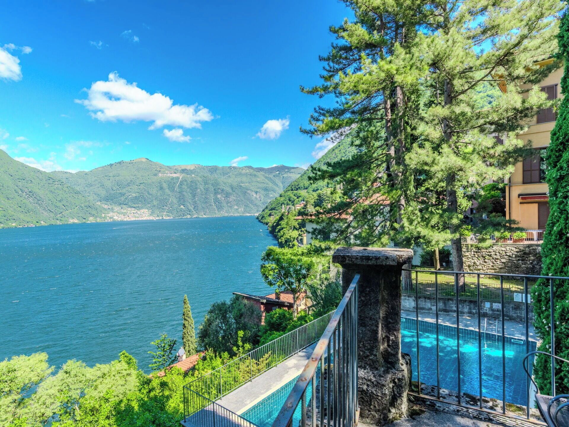 Piscina com estrutura de pedra, cercada por vegetação e com vista para um lago e montanhas ao fundo.