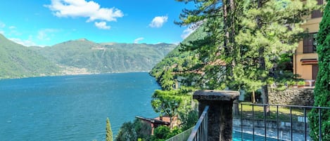 A pool with a stone structure, surrounded by greenery and overlooking a lake with mountains in the background.