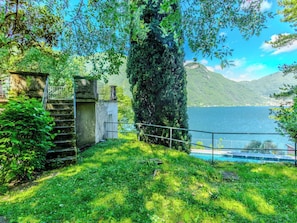 Eine friedliche Seebucht mit einer Steintreppe, die zu einem kleinen Gebäude führt, umgeben von üppiger Vegetation und freiem Blick auf den See und die entfernten Berge.