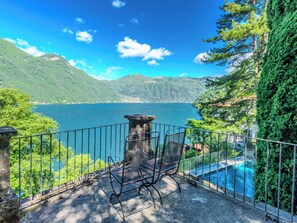 Eine Terrasse mit Steintisch und Stühlen mit Blick auf einen See, umgeben von Grünflächen und Bergen.
