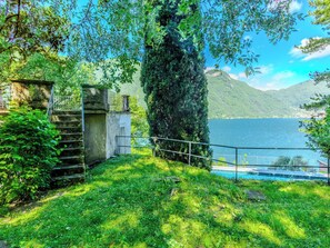 Eine friedliche Seebucht mit einer Steintreppe, die zu einem kleinen Gebäude führt, umgeben von üppiger Vegetation und freiem Blick auf den See und die entfernten Berge.