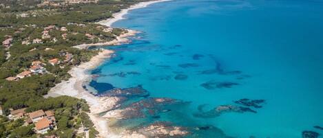 Une vue aérienne d’une zone côtière avec une plage, des eaux d’un bleu limpide et quelques bâtiments dispersés.