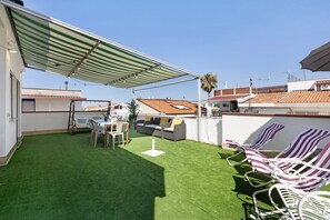 Terrassa al terrat amb gespa artificial, una taula amb cadires, un sofà i gandules amb ratlles.