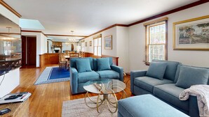 Ein Wohnzimmer mit einem blauen Sofa, einem Glastisch, Holzböden und einem Essbereich im Hintergrund.