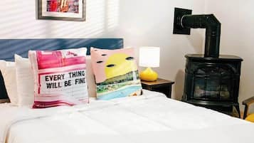 Un lit avec une literie blanche, deux oreillers ornés de motifs, une lampe jaune et un poêle noir.