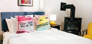 A bed with white bedding, two pillows with text, a yellow lamp, and a black stove. - Stylish studio in central location with fireplace, AC, & tranquil courtyard (Provincetown)