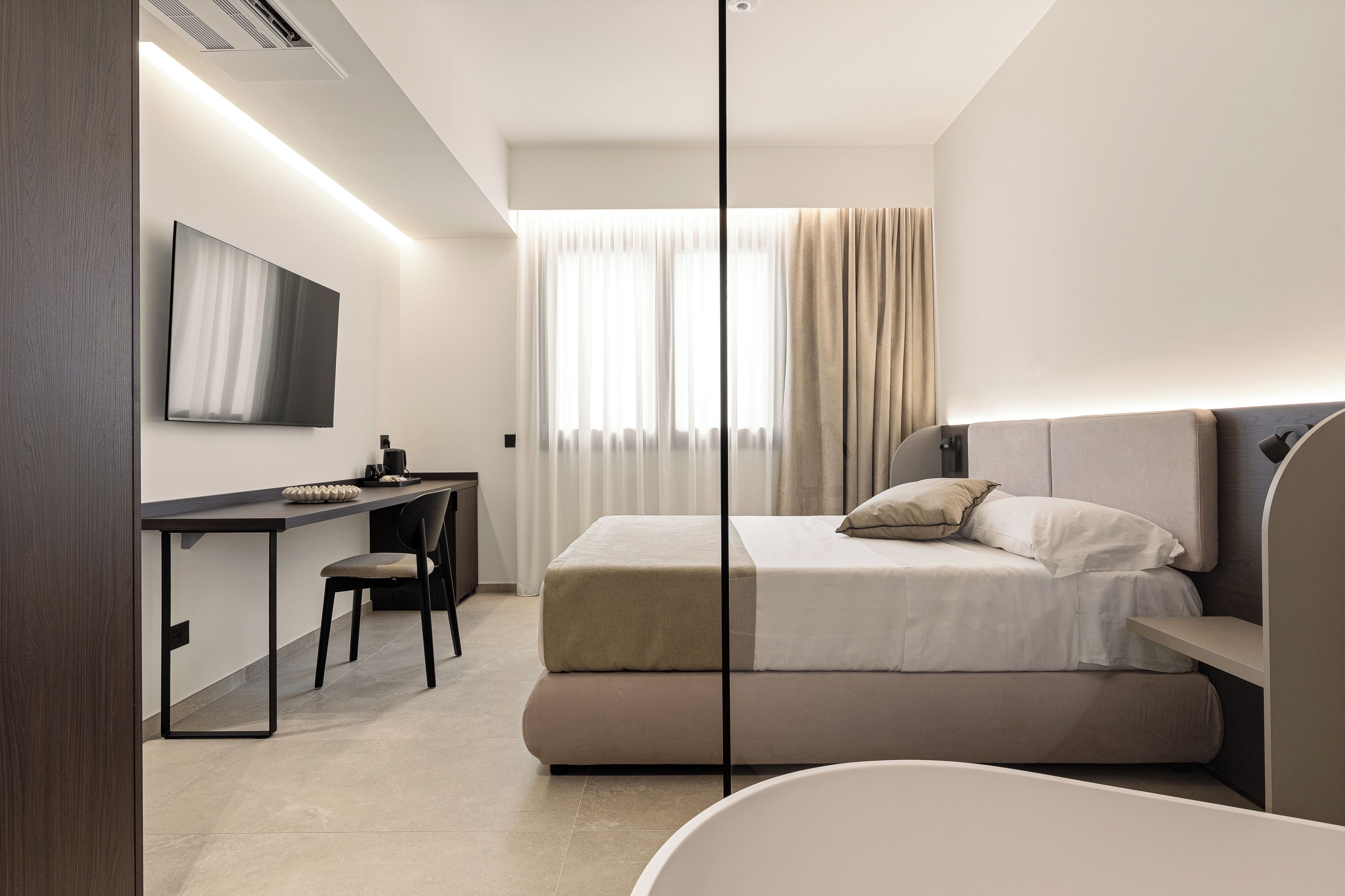A modern hotel room with a bed, desk, chair, and a large window with curtains.