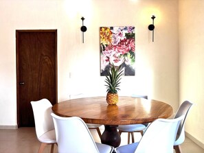 A dining area with a round wooden table, white chairs, a pineapple centerpiece, and a floral painting on the wall. - 'GRAND OPENING' PRICE - FOR RENT - BUCERÍAS 2 BED OASIS WITH PRIVATE POOL 👙 (Bucerías)