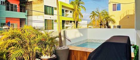 A rooftop pool area with potted plants, a covered seating area, and modern buildings in the background.