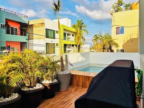 Uma área de piscina no terraço com plantas em vasos, um espaço de estar coberto e edifícios modernos ao fundo.