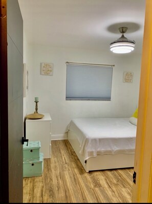 Ein Schlafzimmer mit einem Bett, einem Fenster mit Jalousien, einem Deckenventilator und einem kleinen Schreibtisch mit Lampe.