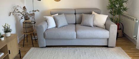 A living room with a grey sofa, a small side table with a plant, a floor lamp, and a large mirror on the wall.