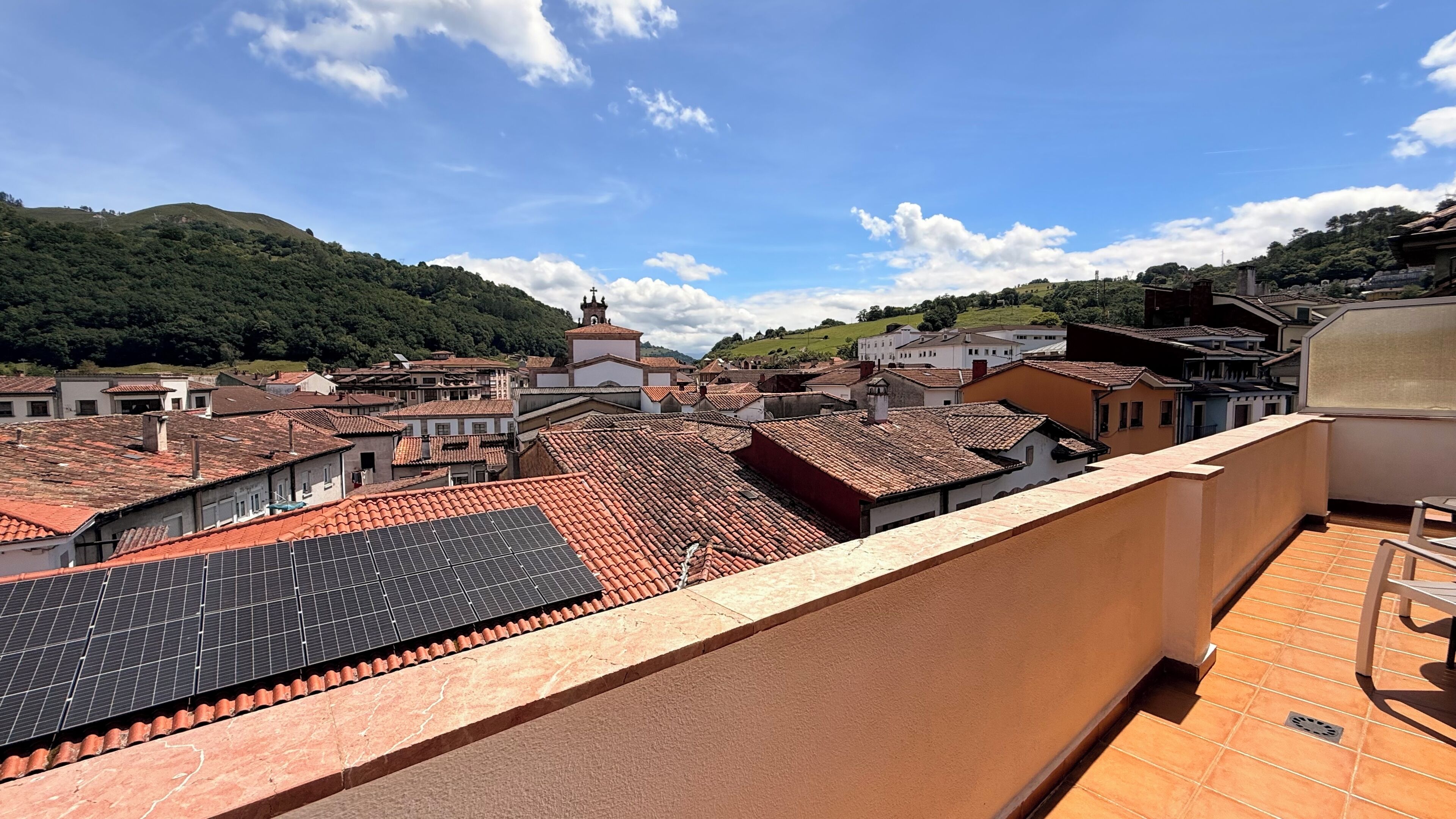 A rooftop with solar panels, terracotta-tiled roofs, and a view of a town with a church.