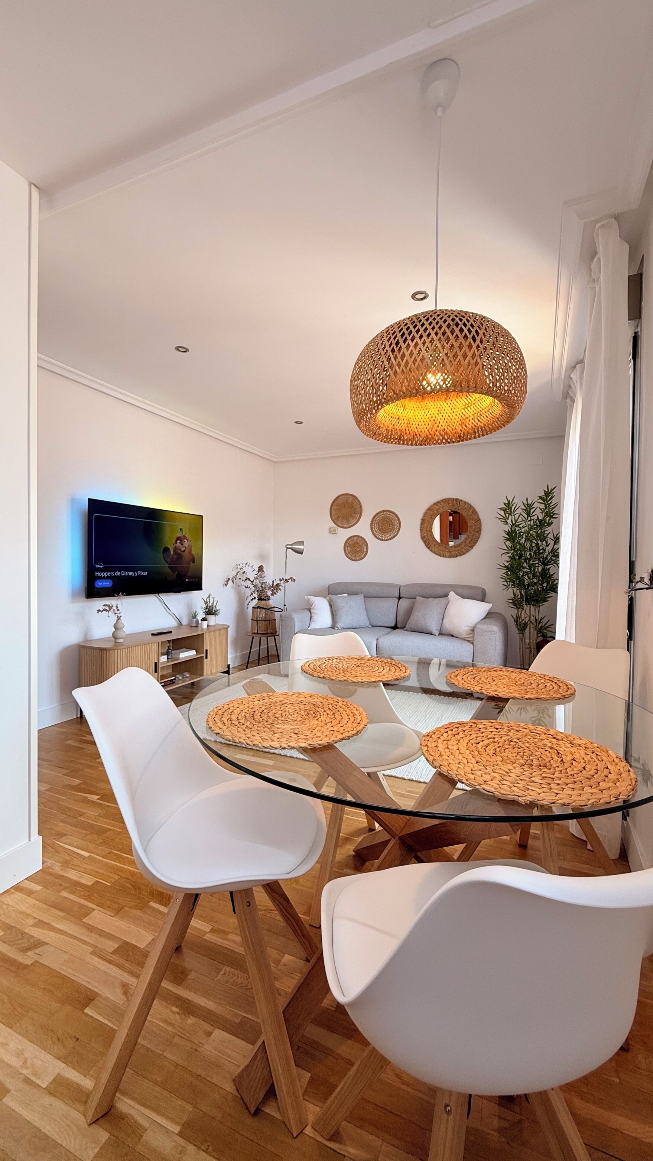 A modern living room with a glass dining table, white chairs, a grey sofa, and a television mounted on the wall.