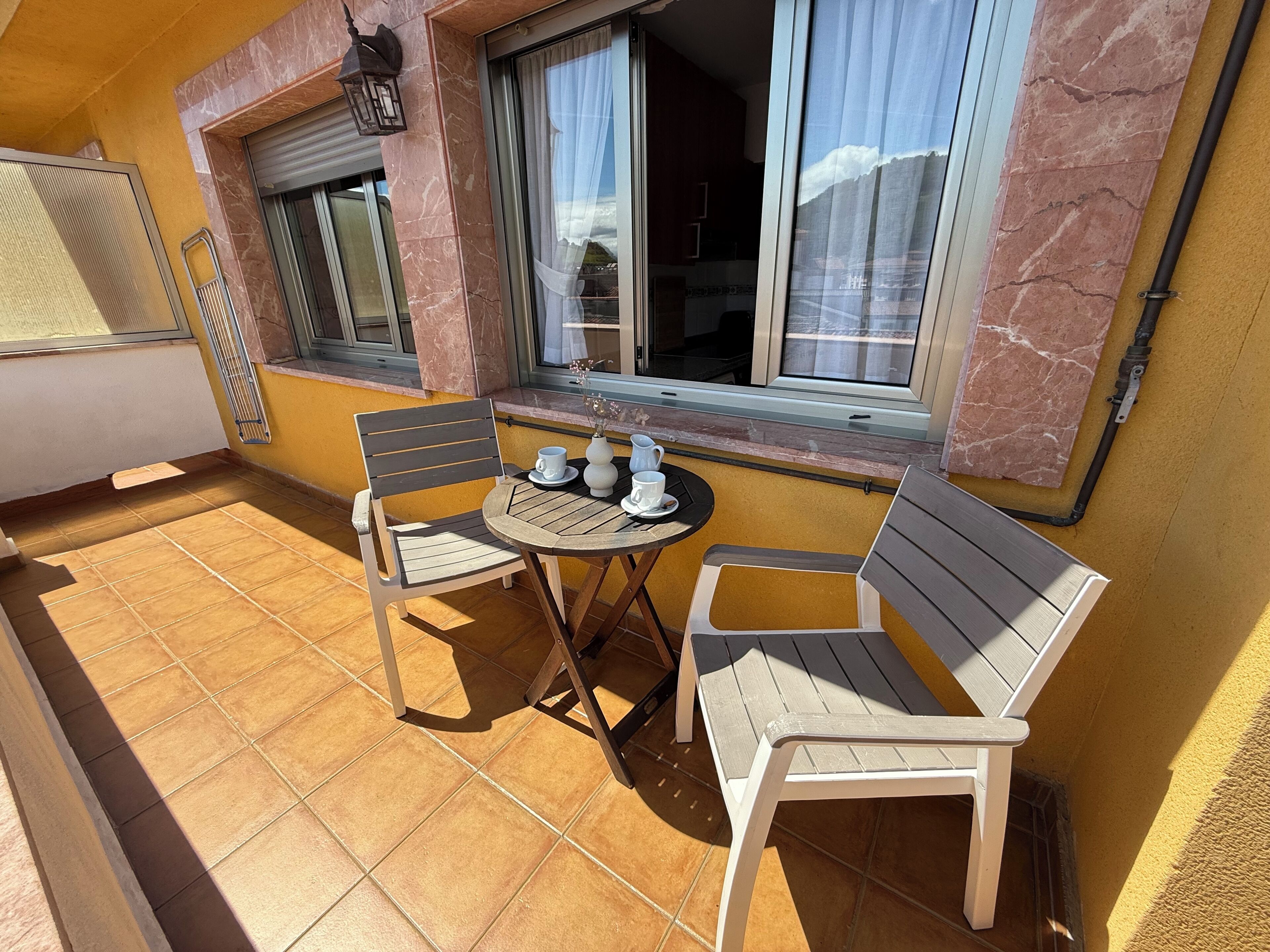 A balcony with a table and two chairs, a window with curtains, and a marble wall. - Hórreo Apartment in downtown Cangas with a terrace and mountain views. (Cangas de Onís)