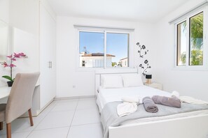 Un dormitorio moderno con una cama, un escritorio, una silla y una ventana con vistas a edificios y árboles.